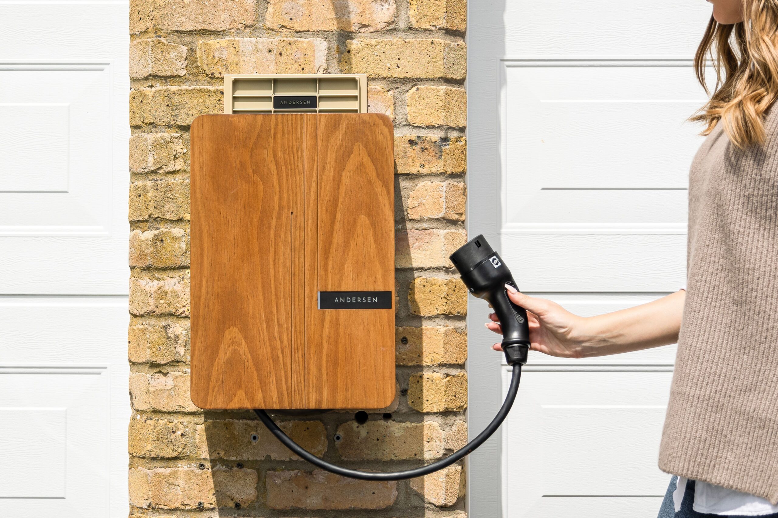Person holding a black power tool next to a brick exterior with a wooden panel and an Andersen mailbox mounted on the wall.