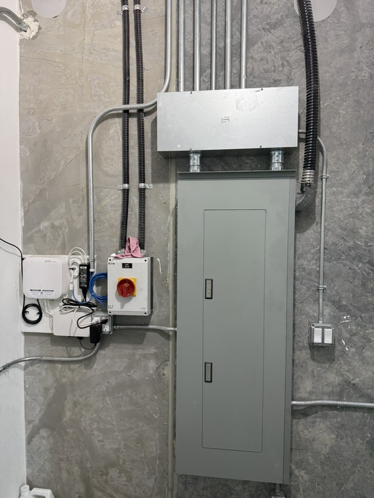 281E135A-177E-410B-896F-0E4D4639F1F0_1_105_c - Kemp Co Electric Industrial electrical panel on a concrete wall with multiple metal conduits; a tall gray cabinet with a door and a red emergency stop box to the left.