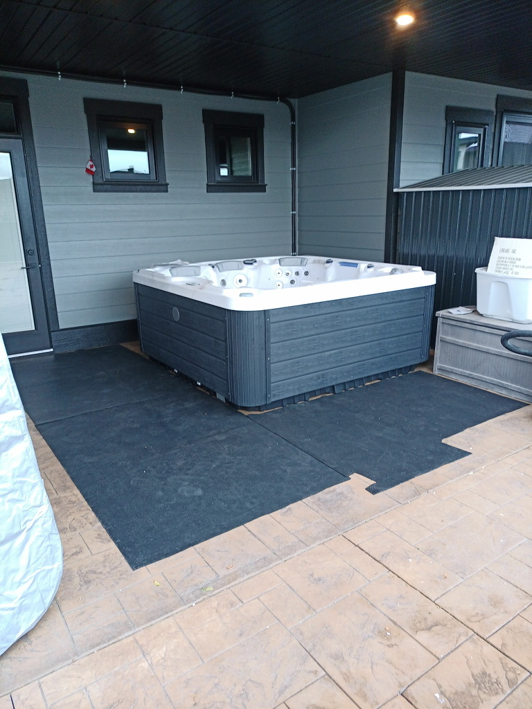 Outdoor hot tub on a covered patio beside a house with gray siding and dark-framed windows, a small Canada flag nearby.