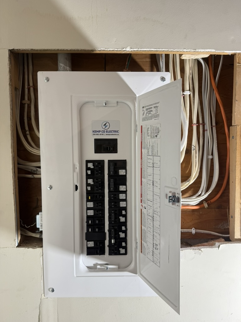 Open electrical panel on a wall with numerous white and orange cables in the background; panel cover tilted to the side revealing circuit breakers inside.