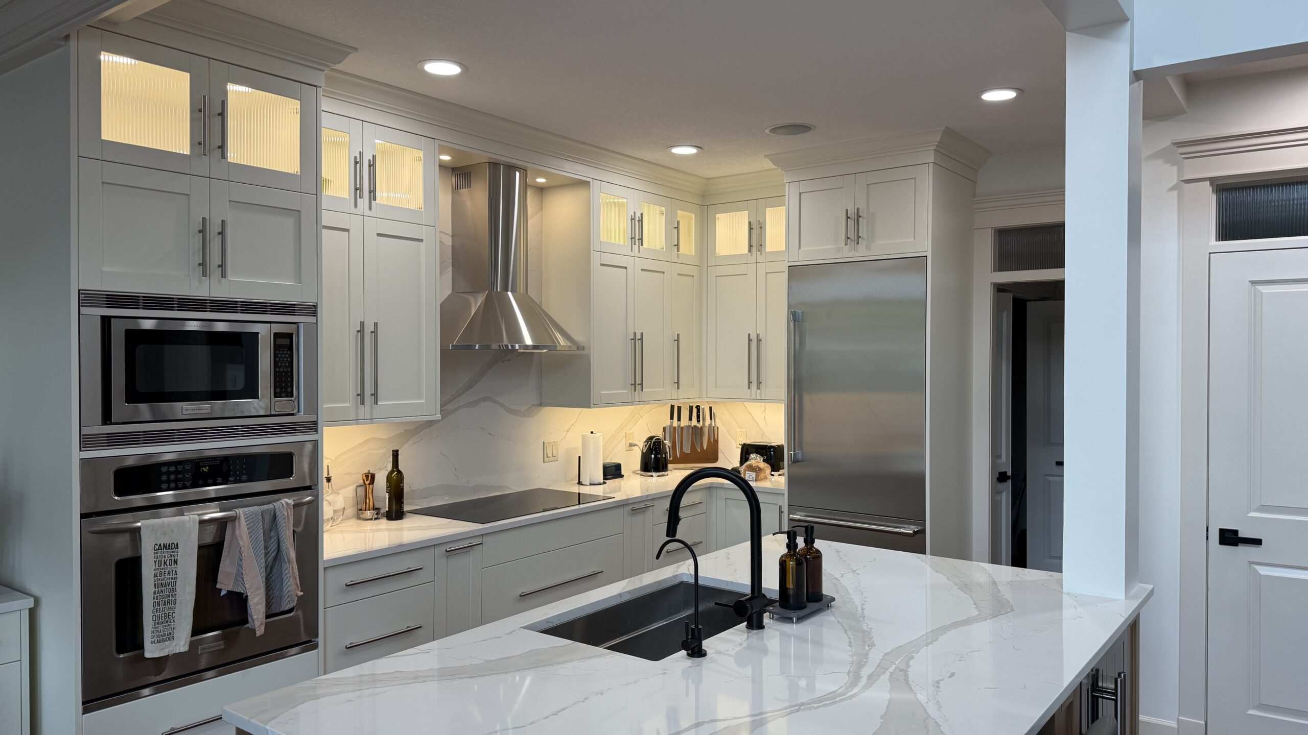 White modern kitchen with marble countertops, island sink, and stainless steel appliances including double ovens and a range hood.