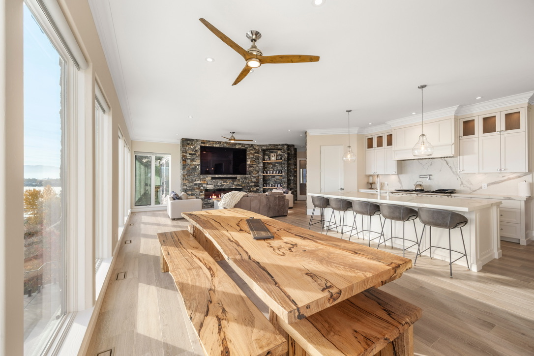 Open-concept living area with a rustic live-edge wooden dining table in the foreground, white kitchen, and a stone fireplace with a TV in the background.