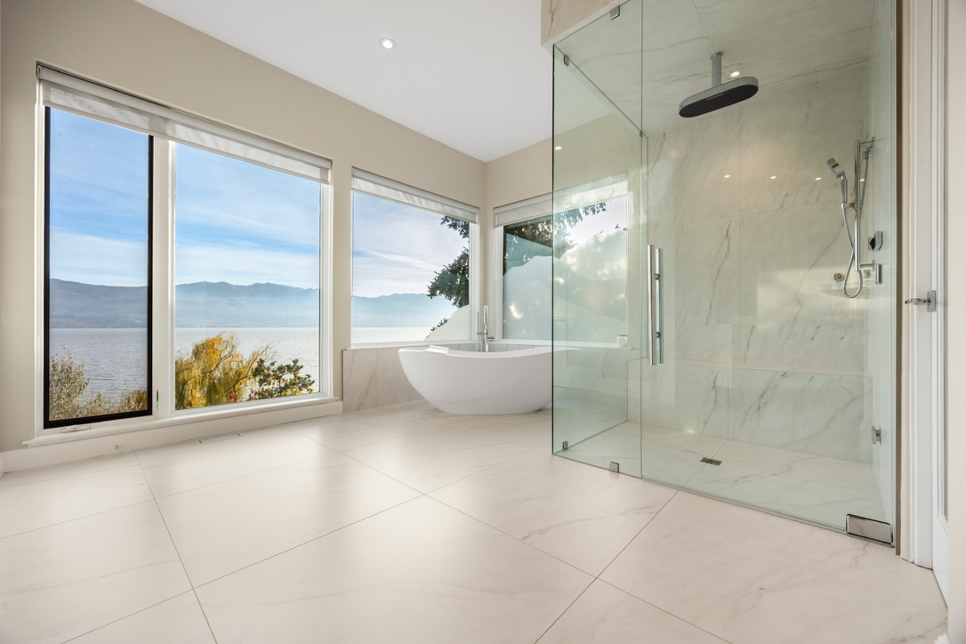 Bright, modern bathroom with large windows overlooking a lake and mountains, a white freestanding tub, and a glass-enclosed marble shower.