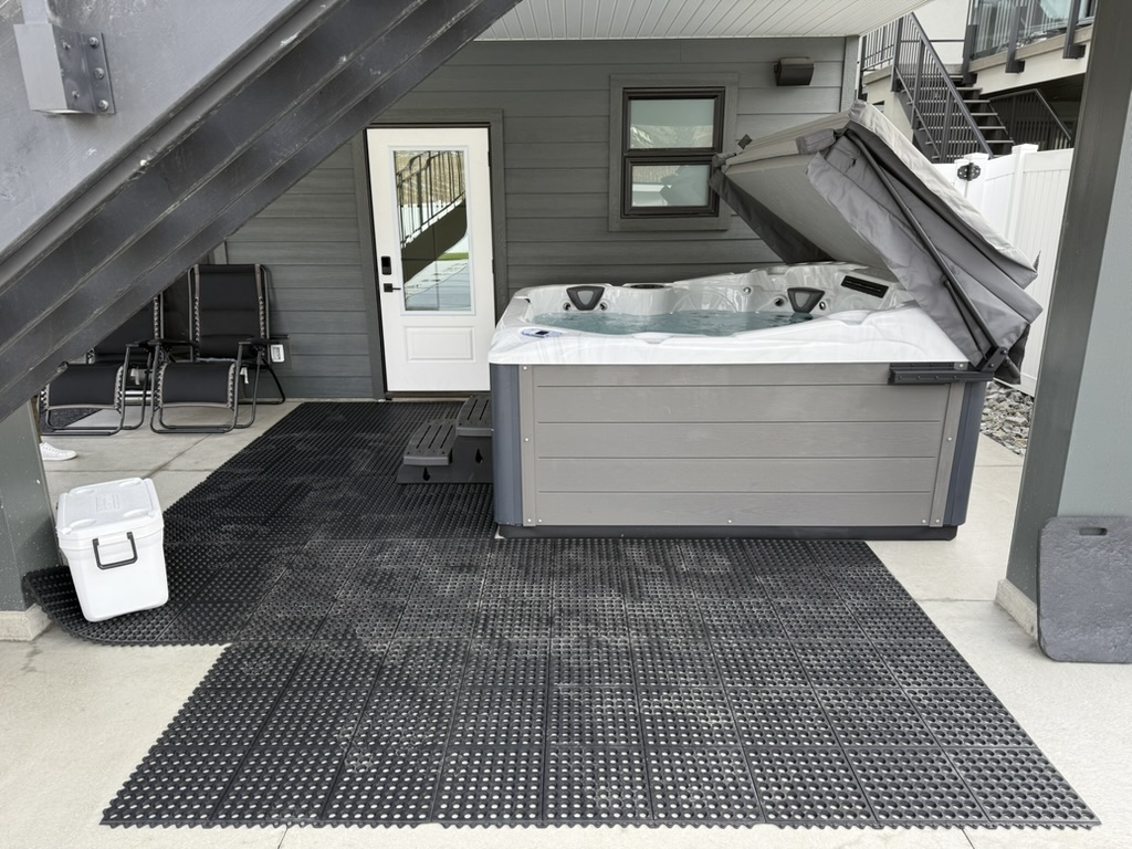 Patio area under a staircase with a gray hot tub, black interlocking mats, a white cooler on the left, and outdoor chairs beside a door in a gray siding home.