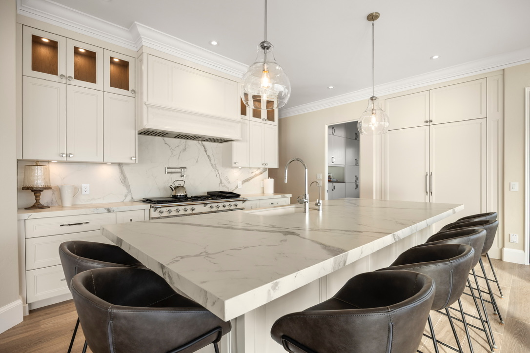 Bright white kitchen with a marble island, stainless range, and pendant lights; six dark leather bar stools along the island.