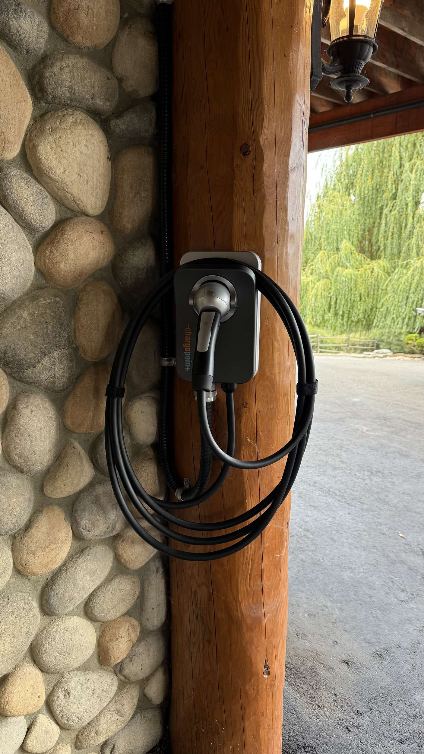 EV charging station mounted on a wooden post with a coiled black cable outdoors beside a stone wall and lantern