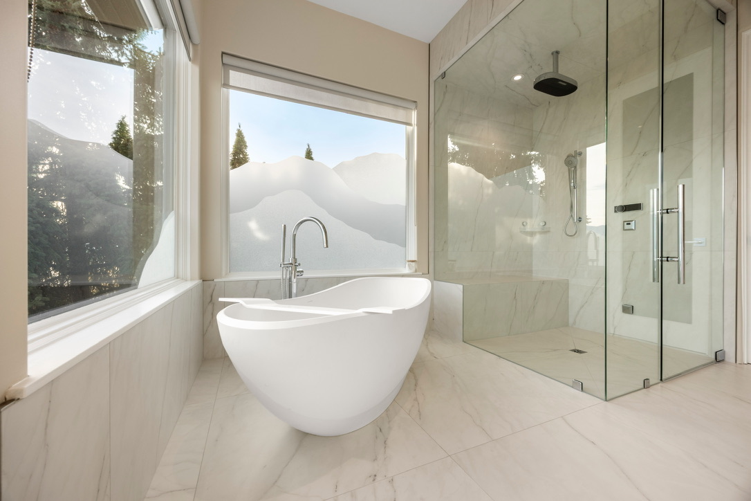 Freestanding white bathtub by a large window with marble floor, next to a glass-walled shower enclosure.