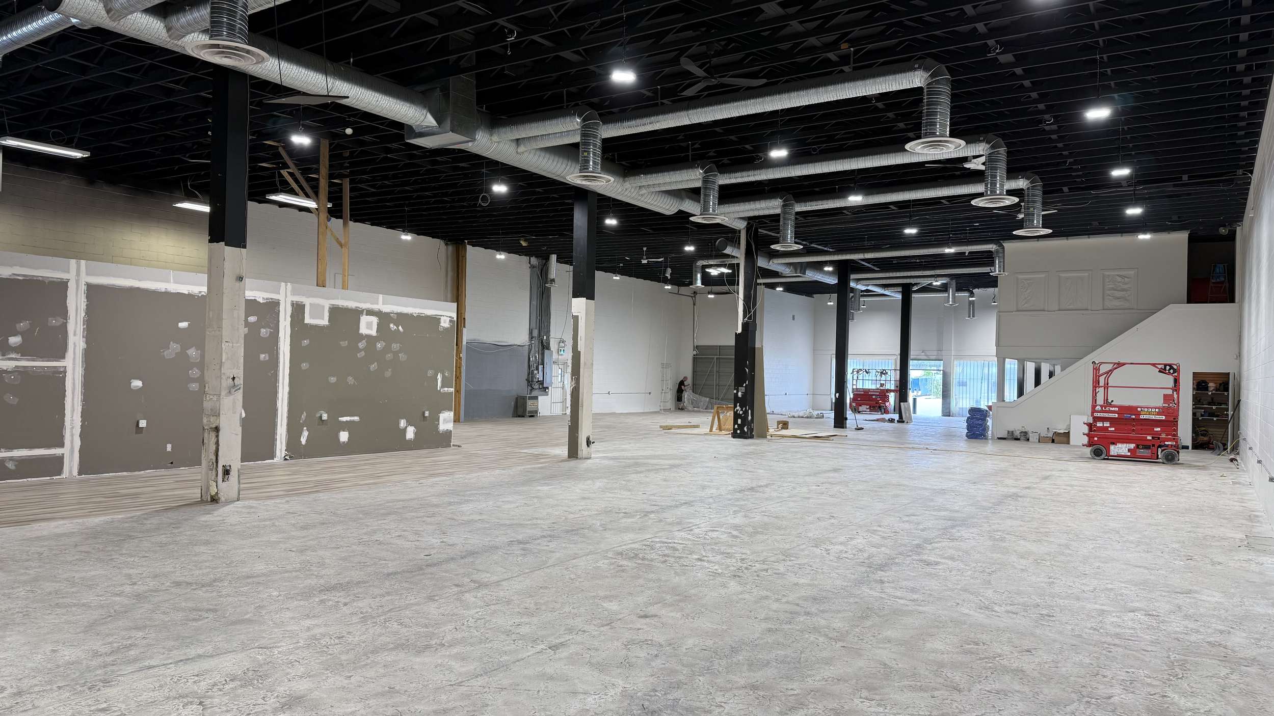 Interior of a large unfinished commercial space with exposed metal ducts and a red scissor lift in the corner on a concrete floor