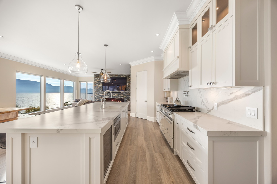 Open-concept kitchen with white cabinets, marble countertops and a large island, overlooking a living area with a stone fireplace and a coastal view through windows.